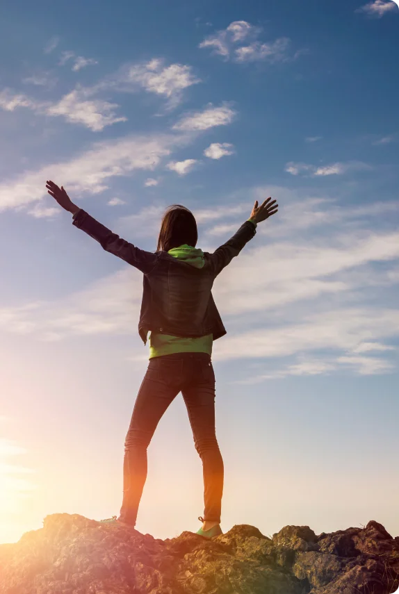 A woman with her back towards the camera is standing on top of a hill. She is raising her arms in triumph. In the background is a blue sky with scattered clouds.