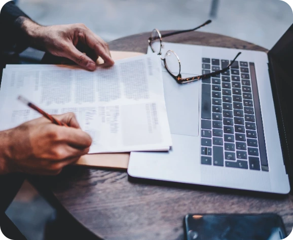A laptop is placed on a table. On the laptop are a pair of glasses. A man's hands are also visible. He is writing with a pencil on a paper document. A partialy obscured mobile phone is beside the laptop..